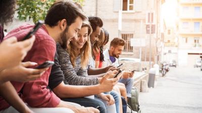 A group of people looking at their mobile phones, sitting on a wall in a sunny city.