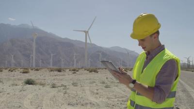 A man wearing a high vis jacket can be seen holding a tablet in front of a wind farm.