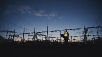 Male Electrical Engineer Using Laptop On Silhouette Power Plant At Twilight