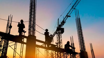 Silhouette of construction workers on scaffolding at sunset, with crane visible against blue-orange sky.