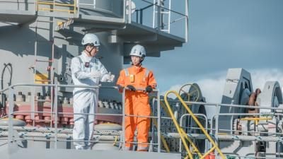 Two maritime workers on ship deck wearing safety gear; one in white coveralls and one in orange.