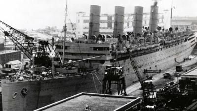 Historical black and white photograph of an ocean liner in dry dock with four prominent smokestacks and shipyard cranes alongside.