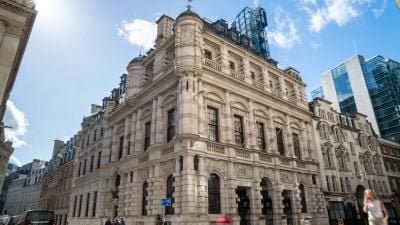 Historic ornate stone building with dome and weathervane in London, modern glass buildings nearby.