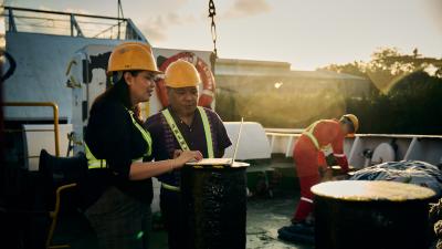 Workers in yellow hard hats and safety vests examining materials on a ship deck at sunset.