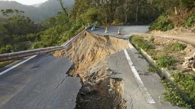 Road collapsed due to landslide on mountainside, with safety cones marking damaged area.