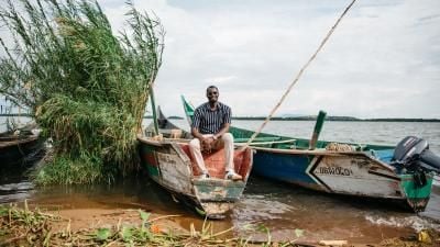 Person sitting on wooden fishing boat at lakeshore with tall reeds and cloudy sky.