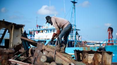 Worker dismantling old boat parts at harbor with blue fishing vessel in background.