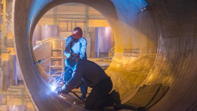 Workers welders inside a large industrial pipe, illuminated by bright welding sparks in a factory setting.