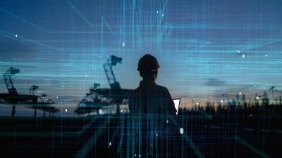 A silhouetted figure wearing a hard hat looks at a device against an industrial backdrop with cranes, overlaid with blue digital grid patterns.