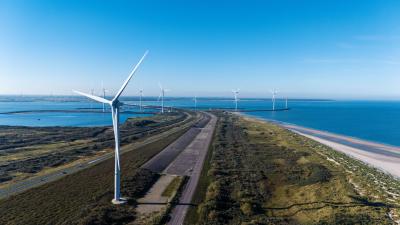 Offshore wind farm with turbines along a coastal road, vibrant blue ocean on both sides.