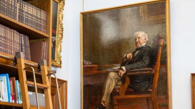 Portrait painting hanging in a library, depicting elderly gentleman in formal attire seated at desk