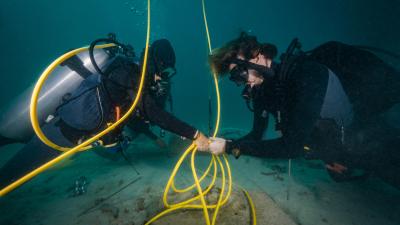 Two scuba divers working underwater with yellow hoses and diving equipment on sandy seafloor.