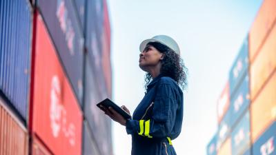 Worker in hard hat and safety gear checking cargo containers at shipping yard.