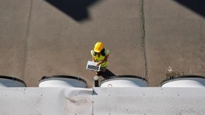 Construction worker in yellow hard hat and safety vest using laptop near industrial tanks.