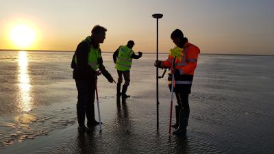 Survey team in high-visibility vests working with measuring equipment on a beach at sunset.