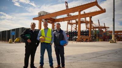 Three workers in safety gear standing at a shipping port with large orange container cranes in the background.