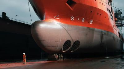 Large orange and gray vessel in dry dock with bulbous bow visible, worker in safety gear standing nearby.