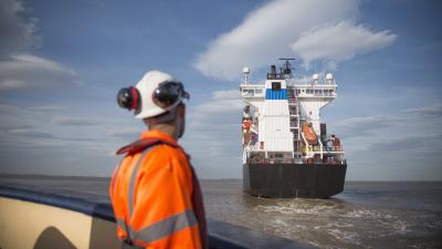 Maritime worker in orange safety gear observing a cargo ship at sea under blue sky.