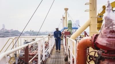 Worker in blue coveralls walking on a metal walkway of an industrial ship with city skyline in background.