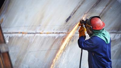 Worker in red hard hat and blue coveralls using angle grinder, creating sparks on metal surface.