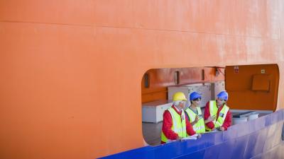 Workers in safety gear and hard hats examining a large orange ship structure through an opening.