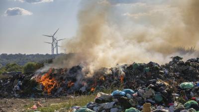 Burning waste dump with thick smoke rising, wind turbines visible in background landscape.