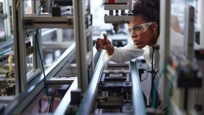 A scientist in protective eyewear examining industrial equipment in a laboratory, focusing on mechanical components.