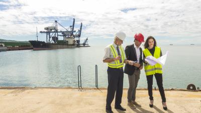 Three professionals reviewing documents at a port, with cargo ship and cranes visible in background.