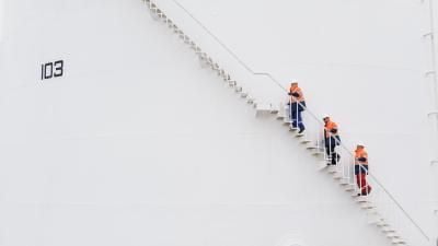 Workers in orange high-visibility clothing ascending a white stairway on what appears to be a large industrial tank numbered 103.