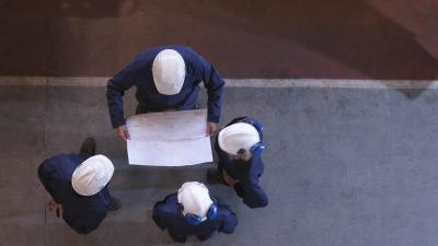 Overhead view of construction workers in blue uniforms and white helmets examining blueprints at a worksite.