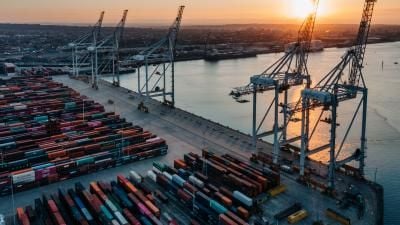 Aerial view of a busy shipping port at sunset with container stacks and large cranes silhouetted against golden light.