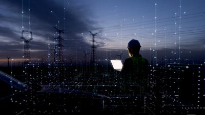 Engineer with hard hat viewing digital overlay of power grid infrastructure at dusk with transmission towers.