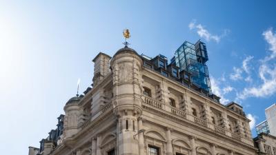 Photograph of the Lloyd's Register Building roofline; a grand Victorian stone building topped with a golden weather vane and a modern glass rooftop extension against a blue sky.