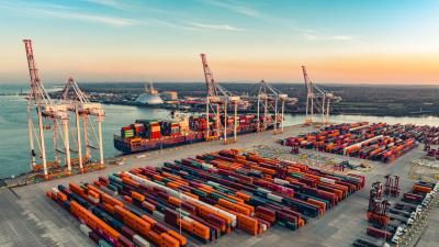 Aerial photograph of a busy container port at sunset, with colourful stacked shipping containers, large cranes, and a cargo vessel docked alongside.