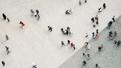 Aerial photograph of people walking across a large grey paved public square.
