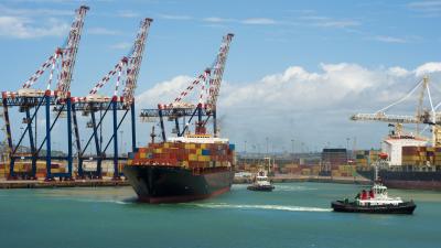 Photograph of a large cargo ship laden with colourful containers, guided by tugboats at a busy port.