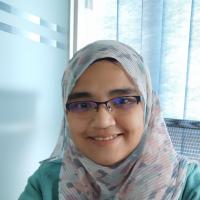 Person wearing glasses and a light-colored patterned hijab, smiling in front of blue curtains.