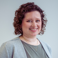 Professional portrait of a person with curly burgundy hair, wearing a gray cardigan and delicate necklaces.