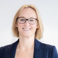 A professional portrait of a person with shoulder-length blonde hair wearing glasses and a navy pinstriped blazer, smiling against a light background.
