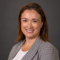 Professional headshot of a smiling woman in a houndstooth blazer and white top against a grey background.