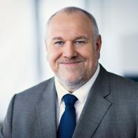 Photograph of a smiling middle-aged man in a grey suit and navy tie, with a blurred office background.