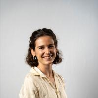 Photograph of a smiling young woman with dark curly hair, wearing a cream silk blouse and hoop earrings.