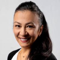 Photograph of a smiling woman with dark hair in a ponytail, wearing a white top, black cardigan, and gold necklaces.